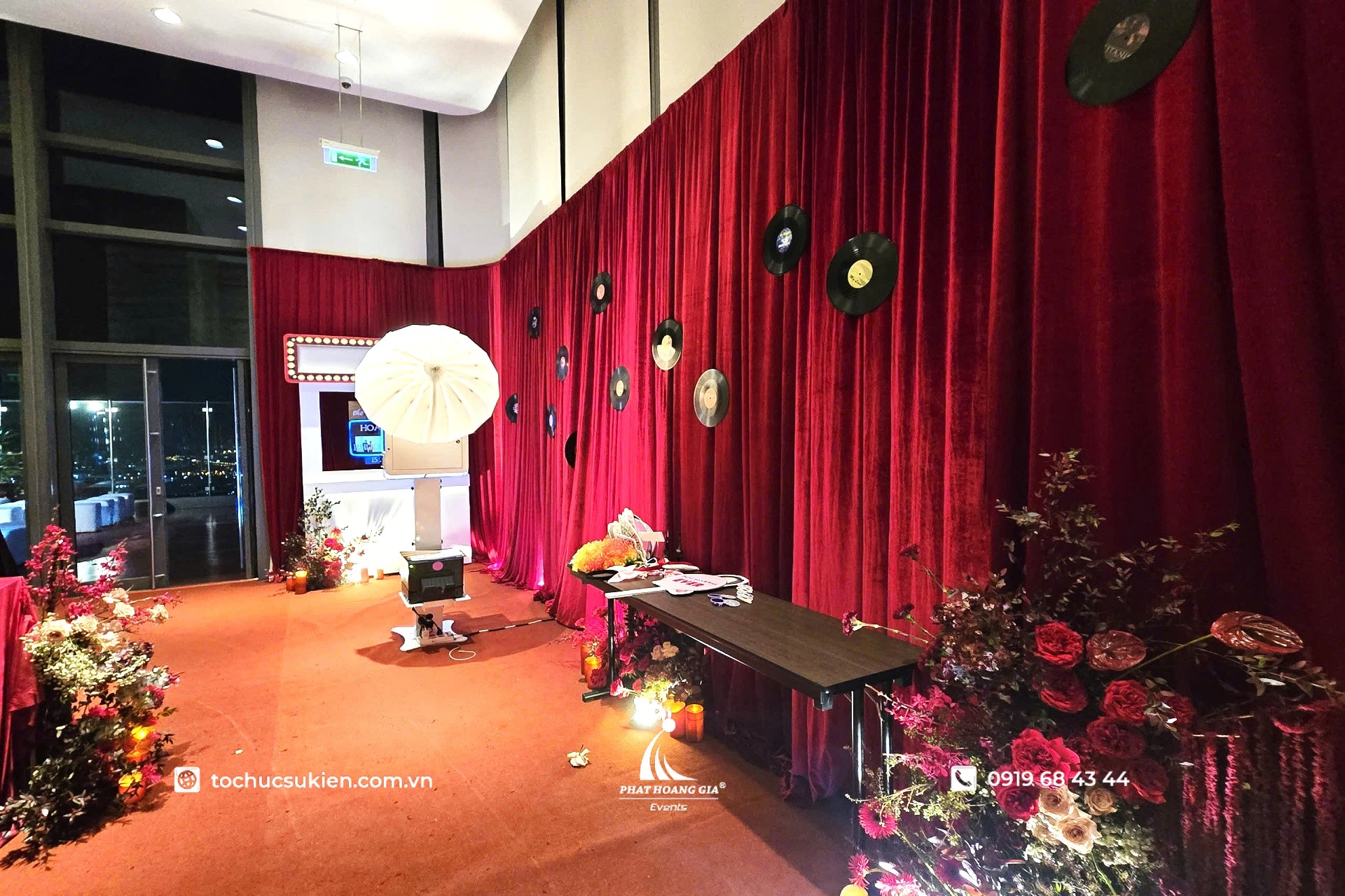 Không gian photobooth sự kiện với backdrop rèm đỏ, booth chụp ảnh, hệ thống đèn chiếu sáng và bàn phụ kiện tương tác cho khách mời