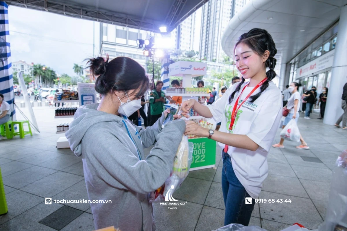 PG phát mẫu dùng thử sản phẩm cho khách hàng trong hoạt động sampling tại booth sự kiện ngoài trời