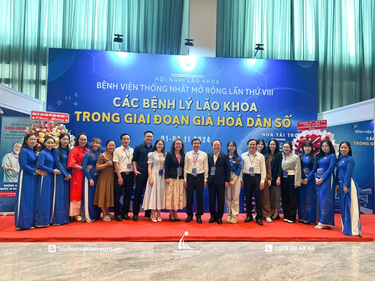 Đoàn đại biểu chụp ảnh lưu niệm trên sân khấu có background hội nghị lão khoa màu xanh nổi bật