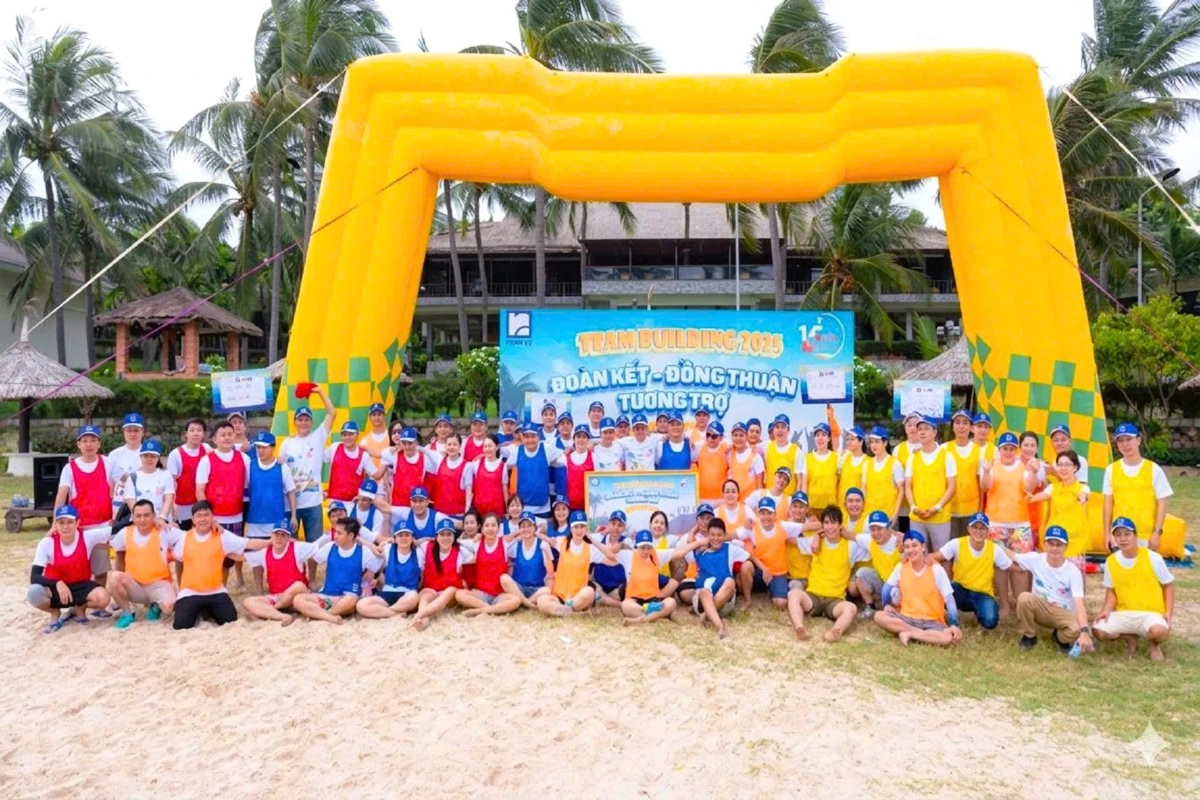 Hình ảnh tập thể chụp ảnh check-in cùng backdrop team building và cổng hơi vàng tại bãi biển.
