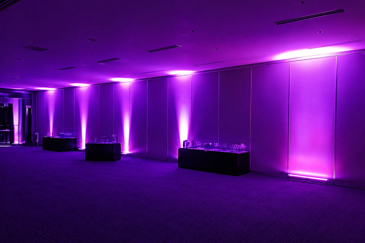 Đèn LED uplight hắt tường màu tím được bố trí dọc không gian sự kiện, tạo hiệu ứng ánh sáng sang trọng và đồng nhất