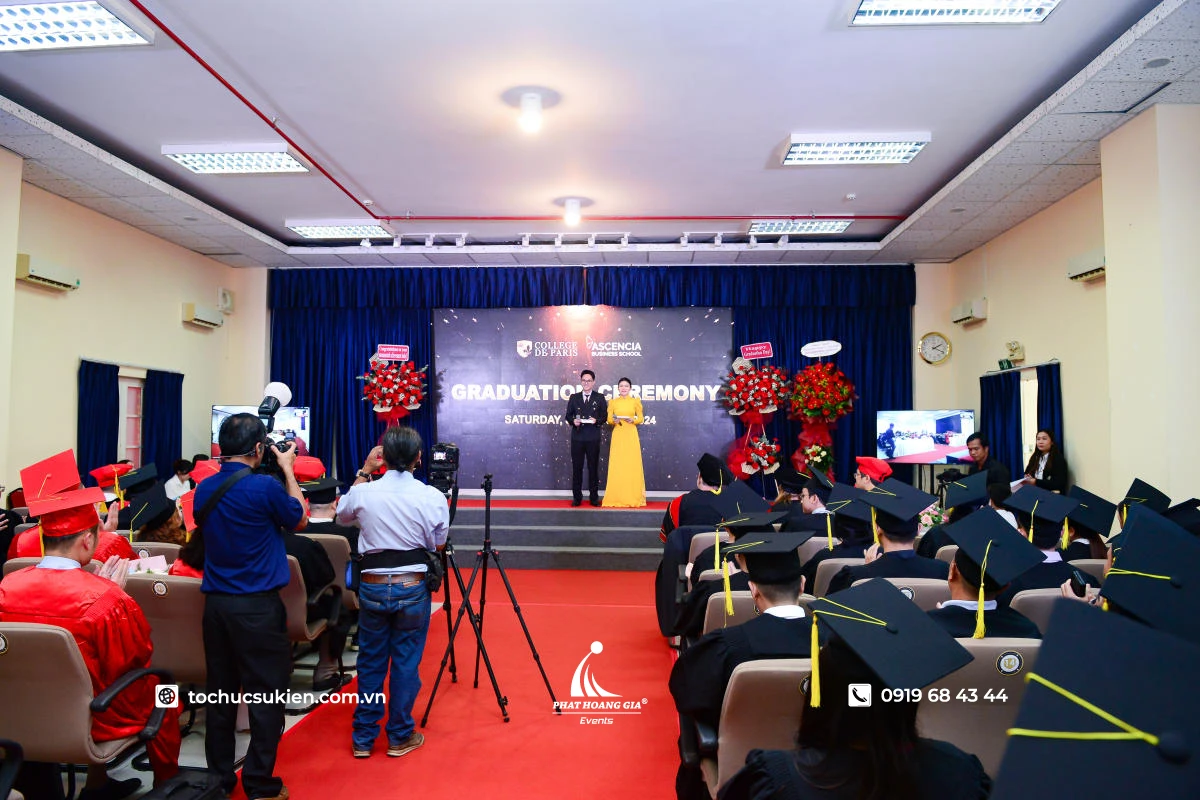 Photographer tác nghiệp tại lễ tốt nghiệp với sân khấu trao bằng và đông đảo sinh viên tham dự trong hội trường