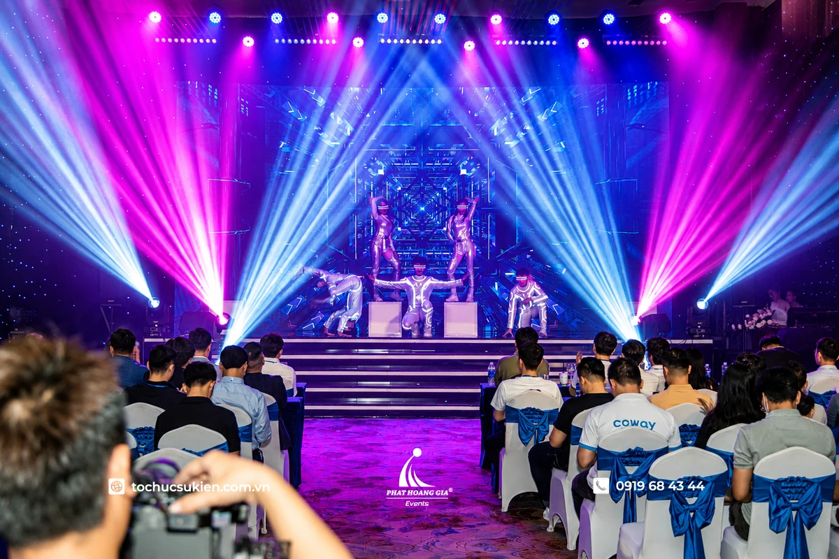 Sân khấu sự kiện trong nhà với màn trình diễn nghệ thuật hiện đại, hệ thống đèn moving head tạo hiệu ứng tia sáng tím xanh nổi bật kết hợp màn hình LED lớn phía sau