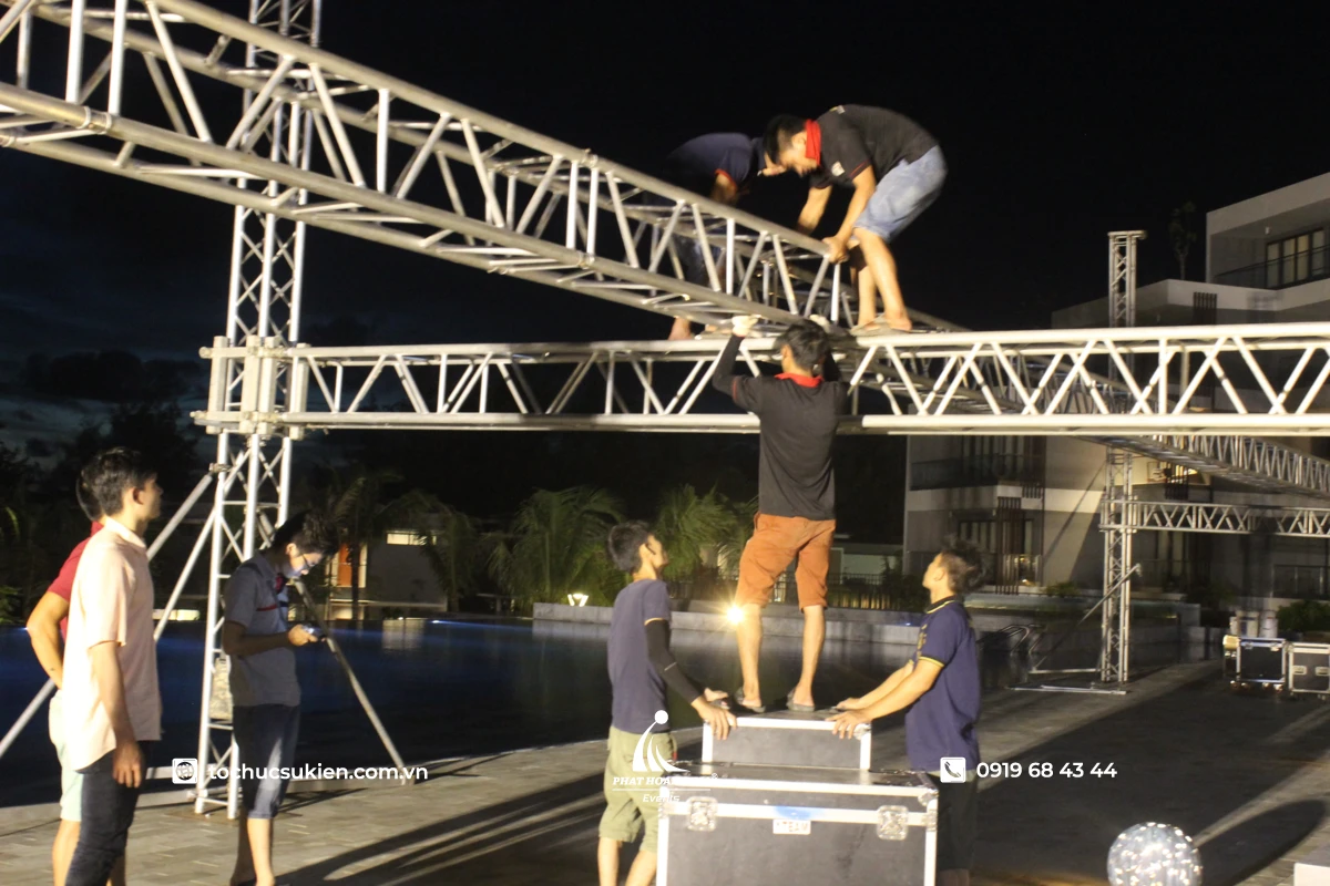 Kỹ thuật viên đang hoàn thiện setup sân khấu ngoài trời với giàn khung truss và hệ thống âm thanh ánh sáng.