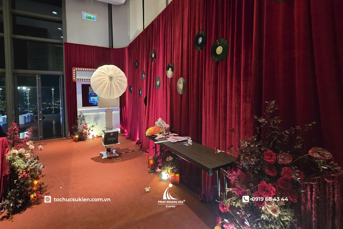 Khu vực setup photo booth trong sự kiện với backdrop rèm đỏ sang trọng, hệ thống đèn chụp ảnh, bàn props tạo dáng và không gian trang trí hoa theo phong cách gala cao cấp