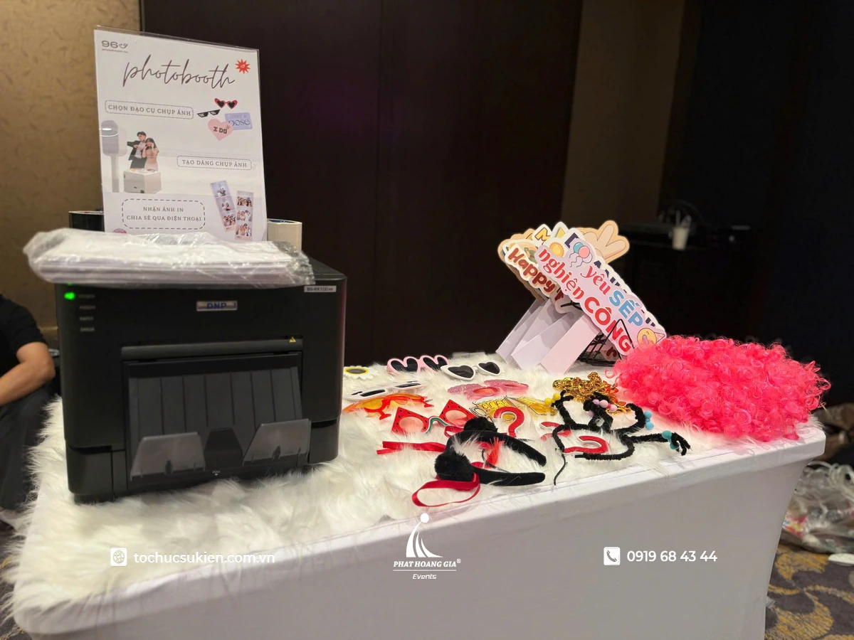 Bàn phụ kiện photo booth với máy in ảnh lấy liền, kính vui nhộn, bảng hashtag cầm tay và đạo cụ tạo dáng dành cho khách tham dự sự kiện doanh nghiệp