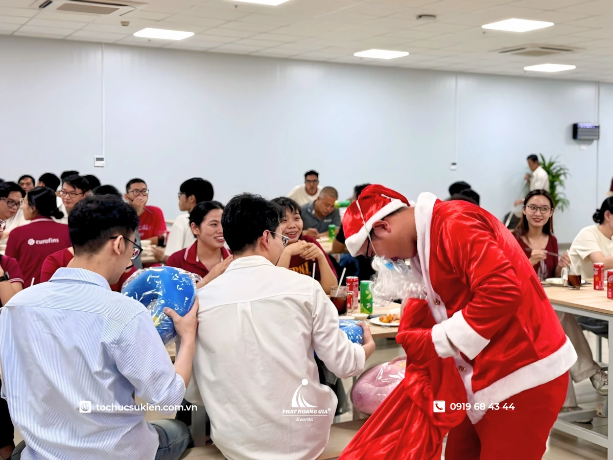 Ông già Noel mở túi phát quà tặng cho tập thể nhân viên đang ngồi quanh bàn ăn trong tiệc giáng sinh