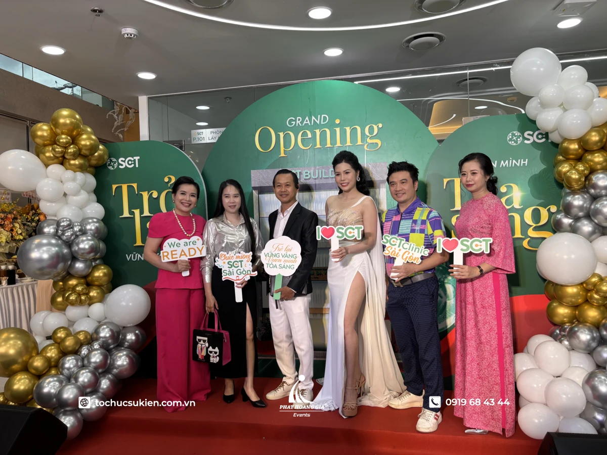Khách mời cầm bảng hashtag chụp ảnh cạnh background khai trương trang trí bóng bay